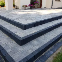 Schody wejściowe wyłożone szarą kostką brukową z ciemnoszarym obramowaniem, prowadzące do domu z jasną elewacją i donicami z kwiatami.