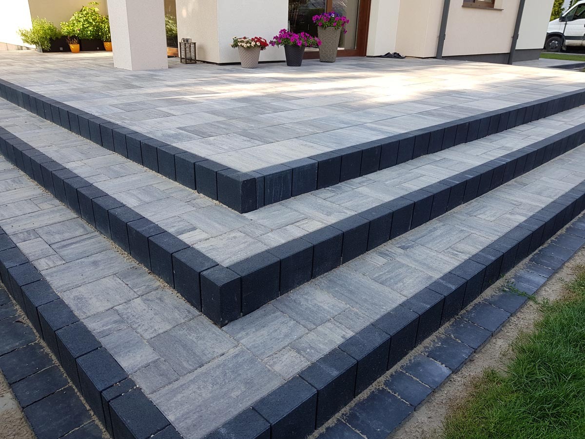 Schody wejściowe wyłożone szarą kostką brukową z ciemnoszarym obramowaniem, prowadzące do domu z jasną elewacją i donicami z kwiatami.