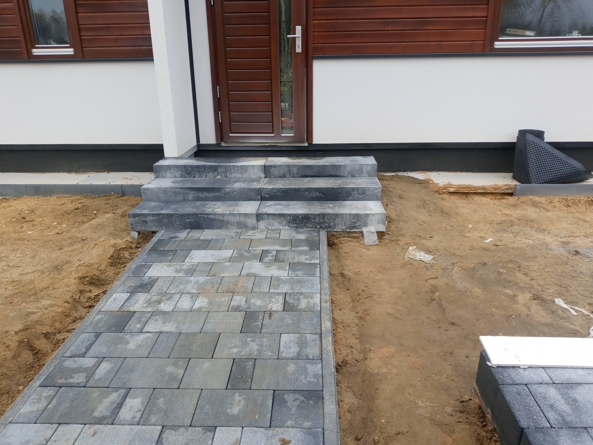 Ułożona ścieżka z szarej kostki brukowej prowadząca do schodów wejściowych domu z brązowymi oknami, widoczne niedokończone prace ziemne po bokach.
