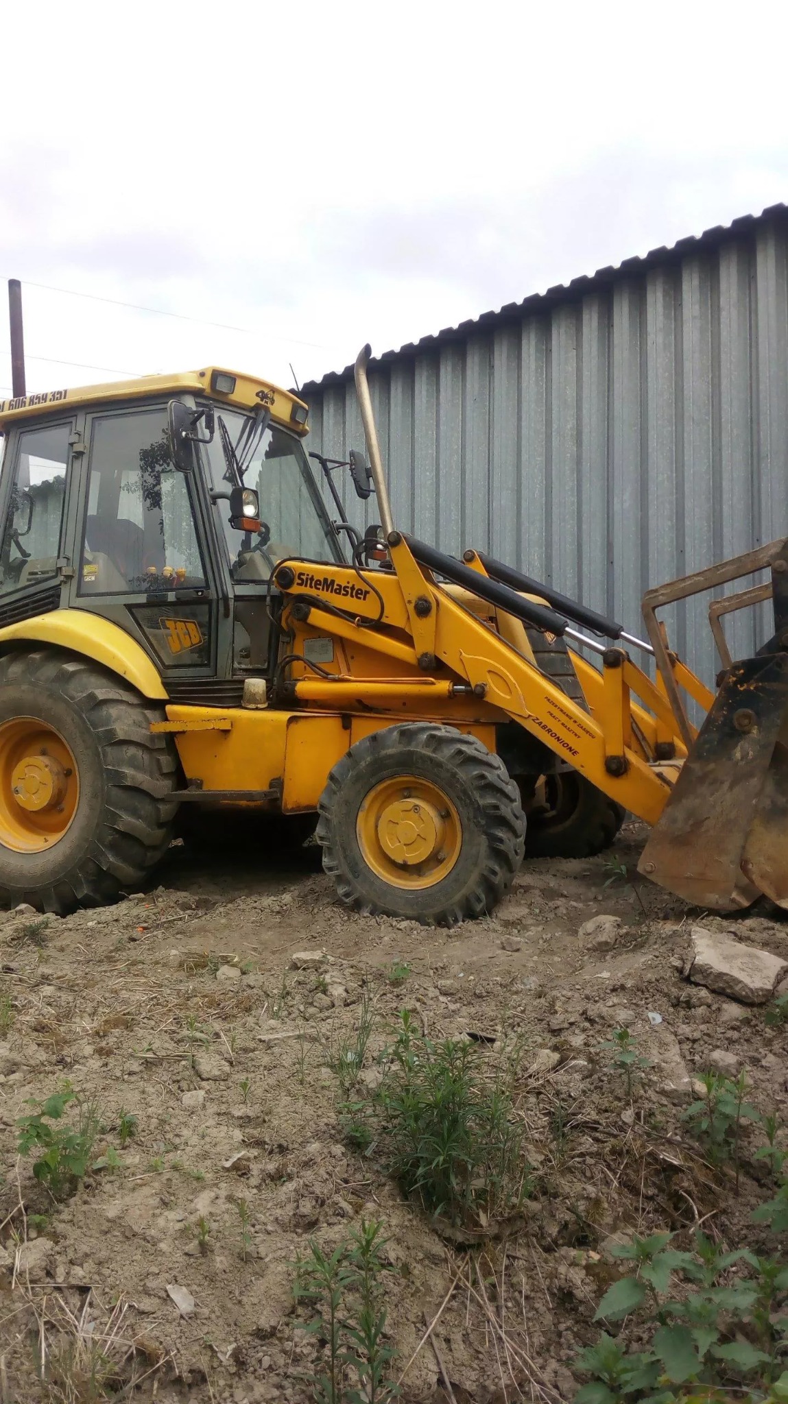 Żółta koparko-ładowarka SiteMaster JCB na placu budowy, zaparkowana obok blaszanej ściany, widoczne ślady użycia i ziemia wokół.