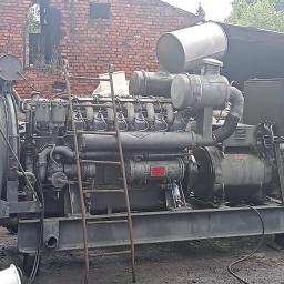 Kręcę Wałek - Duży, szary agregat prądotwórczy z metalową drabinką opartą o bok, stojący na zewnątrz na ziemi, w tle ceglany budynek z dziurami w ścianie.