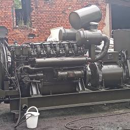 Kręcę Wałek - Duży, oliwkowozielony agregat prądotwórczy z białym wiadrem i wężem na brudnym podłożu, w tle ceglany budynek z pustym oknem.