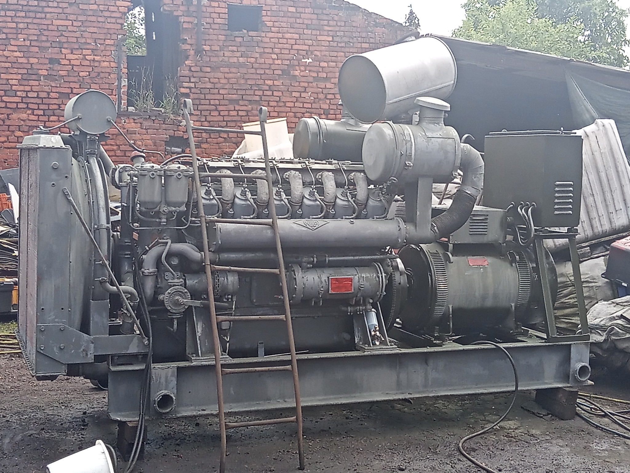 Duży, szary agregat prądotwórczy z metalową drabinką opartą o bok, stojący na zewnątrz na ziemi, w tle ceglany budynek z dziurami w ścianie.