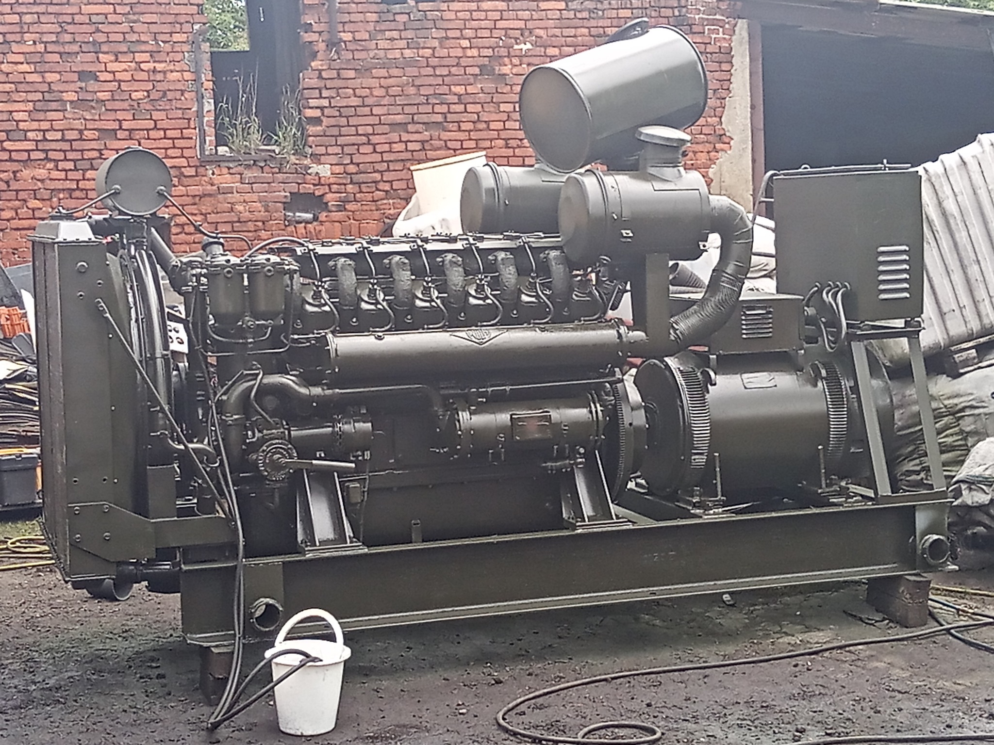 Duży, oliwkowozielony agregat prądotwórczy z białym wiadrem i wężem na brudnym podłożu, w tle ceglany budynek z pustym oknem.