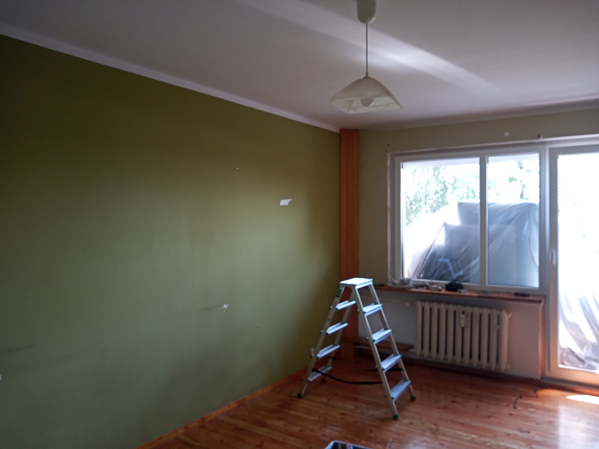 Pokój przed odświeżeniem: oliwkowa ściana z usuniętymi elementami, aluminiowa drabina rozstawiona na drewnianej podłodze, okno zabezpieczone folią malarską.