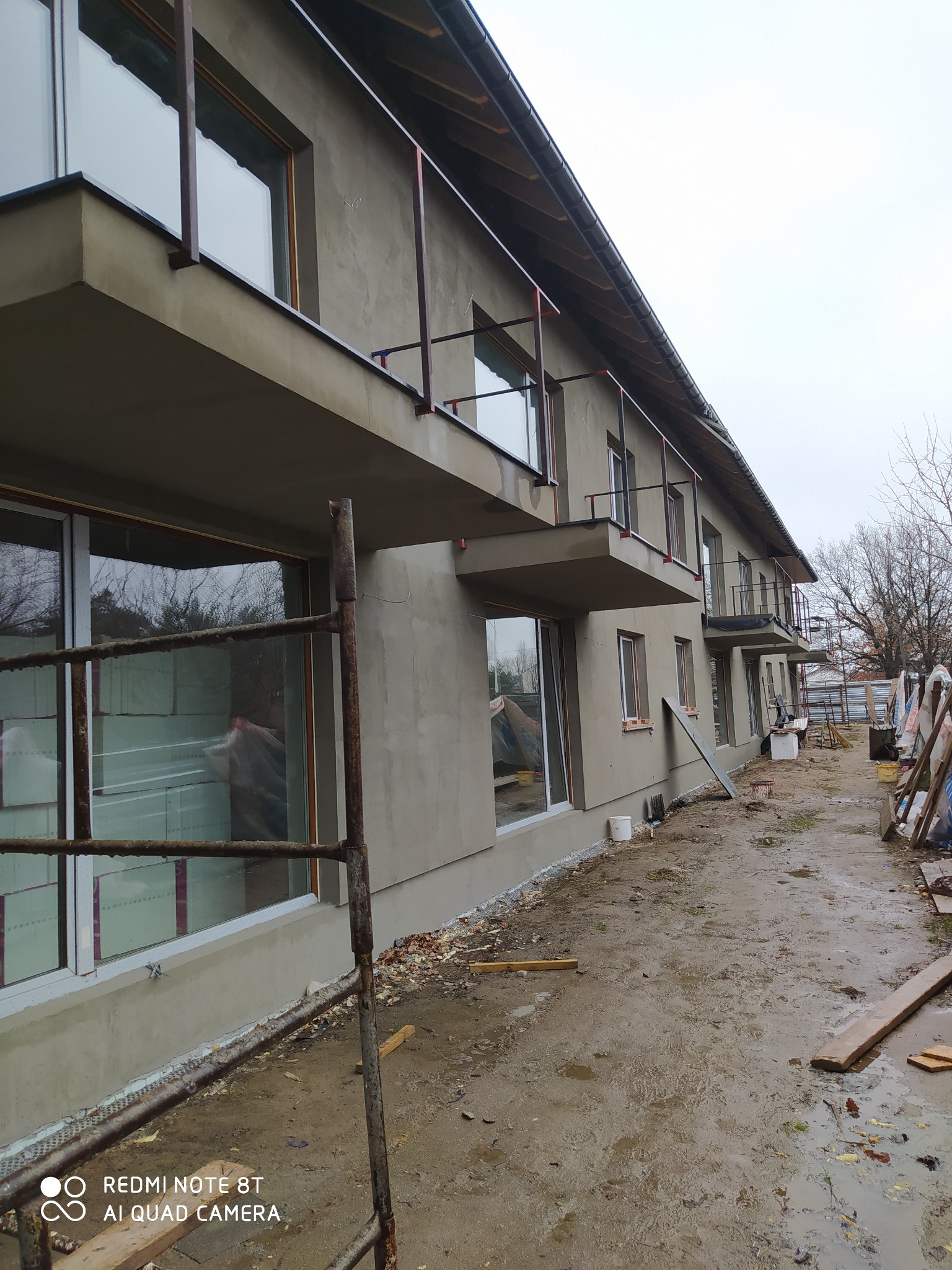 Surowy, dwukondygnacyjny dom murowany w trakcie budowy, z widocznymi rusztowaniami, oknami z izolacją i balkonami z metalowymi barierkami, otoczony błotnistym terenem budowy.