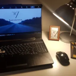 Laptop z podświetloną klawiaturą i ekranem wyświetlającym logo firmy IT, stojący na biurku obok lampki, ramki ze zdjęciem i notatnika z długopisem.
