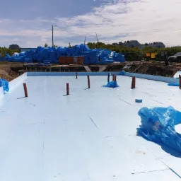 Przygotowanie płyty fundamentowej z izolacją termiczną z polistyrenu ekstrudowanego (XPS), widoczne pionowe rury instalacyjne i zapasy materiału budowlanego w niebieskich opakowaniach na placu budowy.