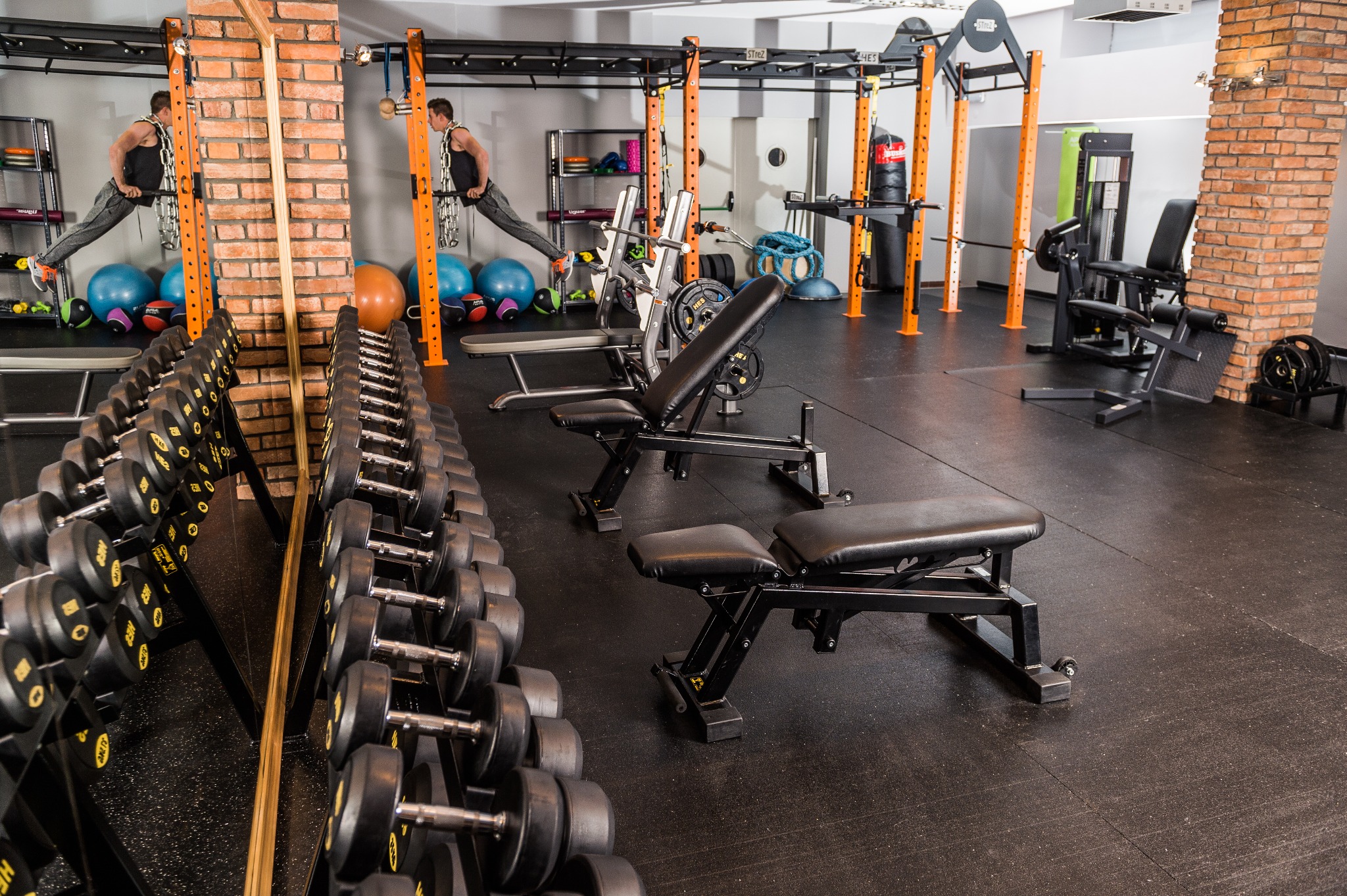 Widok na siłownię z rzędem hantli, ławeczkami do ćwiczeń i ćwiczącym mężczyzną na drążku w tle, odbicie w lustrze