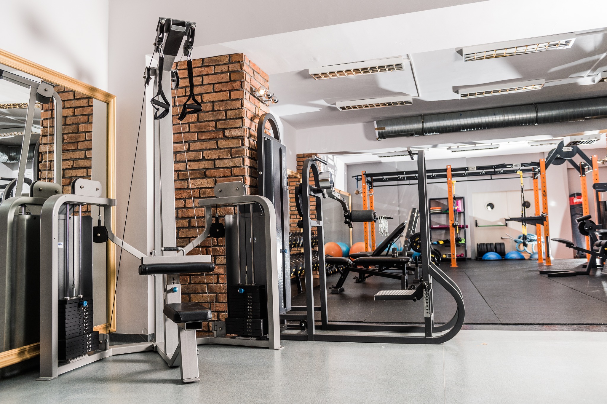 Siłownia z różnorodnym sprzętem do ćwiczeń, w tym maszyny z obciążeniami i drabinki, z ceglanym filarem w tle.