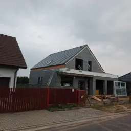 Budowa domu jednorodzinnego w Ostrzeszowie w trakcie montażu pokrycia dachowego z widocznymi oknami dachowymi oraz drabina oparta o ścianę budynku.