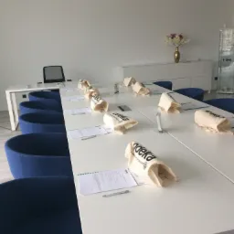 Sala konferencyjna z długim białym stołem, niebieskimi krzesłami, materiałowymi torbami z logo i dokumentami dla uczestników, przygotowana do szkolenia.