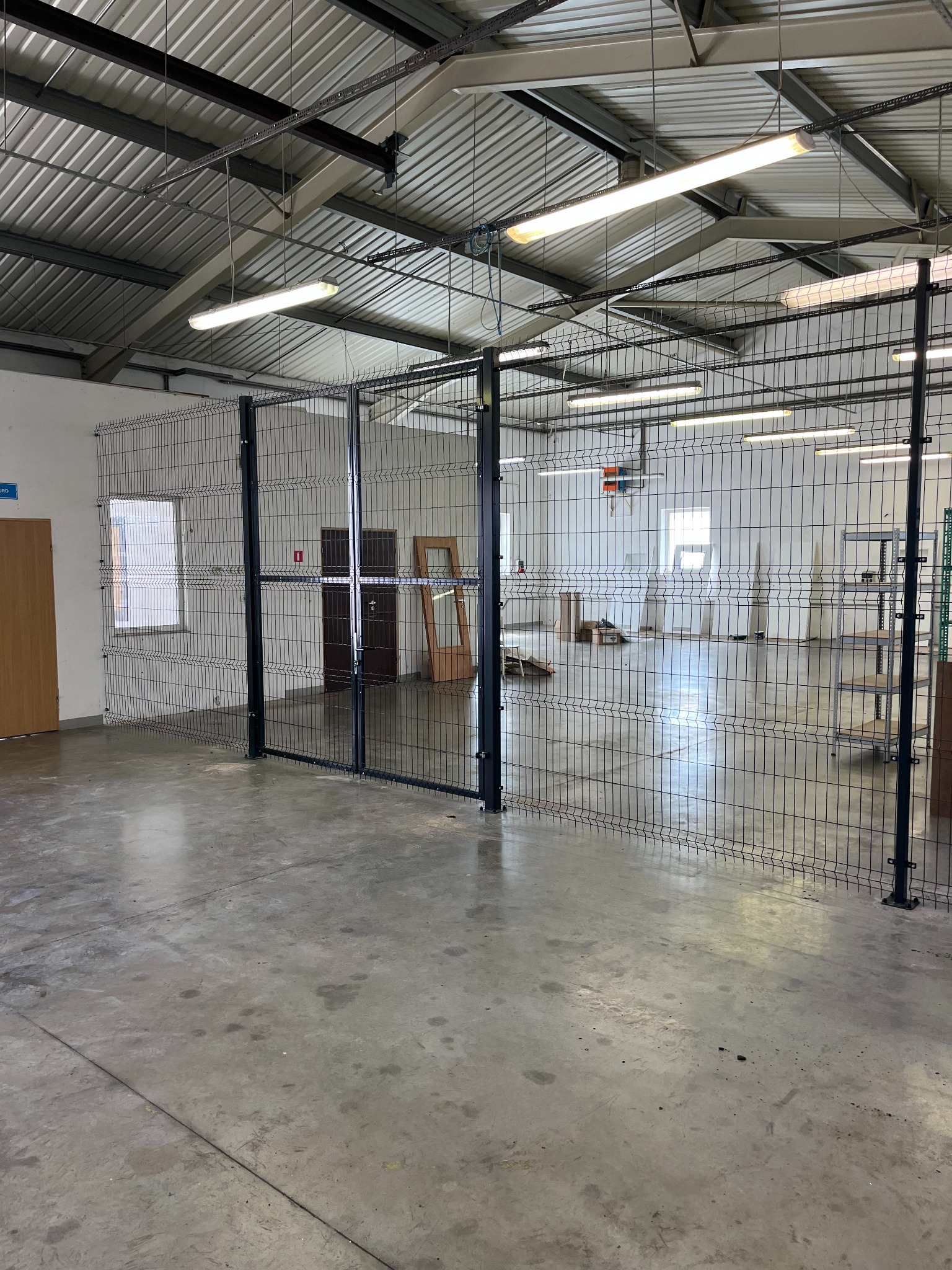 Metalowa siatka ogrodzeniowa w hali magazynowej, z widocznymi elementami konstrukcyjnymi i oświetleniem. Przez siatkę widoczne drzwi i okna.