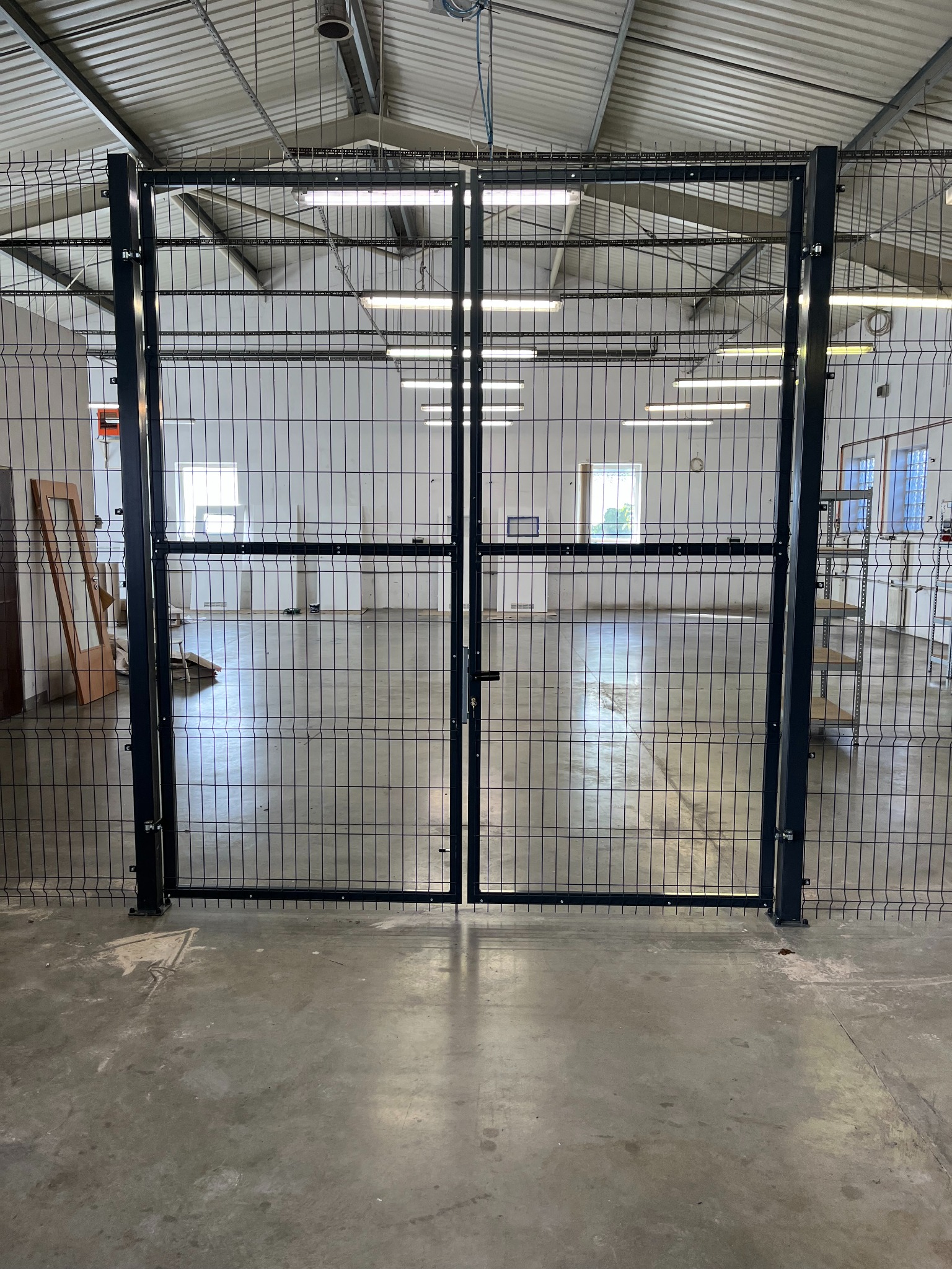 Zamknięta, metalowa brama z siatki w hali przemysłowej. Widoczne mocowania i zamek. W tle okna i oświetlenie hali. Betonowa posadzka.