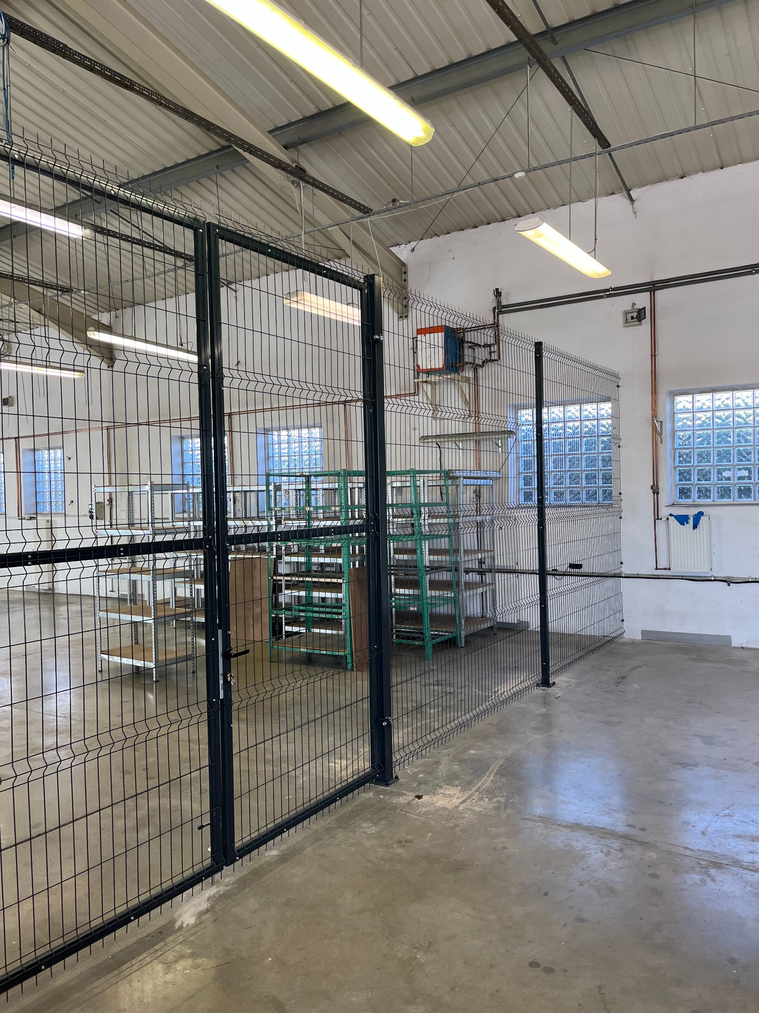 Metalowa siatka ogrodzeniowa z bramą, oddzielająca część hali magazynowej. W tle widoczne regały i okna z luksferami. Oświetlenie jarzeniowe.
