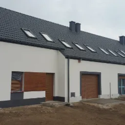 Nowoczesne szeregowe domy z białą elewacją, ciemną stolarką okienną i grafitowym dachem z oknami dachowymi, teren wokół w trakcie zagospodarowywania.