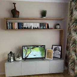 Nowoczesny salon z szarą szafką RTV, telewizorem, drewnianą półką na książki i dekoracjami, oświetlony naturalnym światłem wpadającym przez okno z wzorzystymi zasłonami.