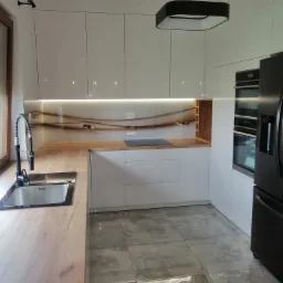 Nowoczesna kuchnia z białymi, błyszczącymi szafkami, drewnianym blatem i szklanym panelem nad blatem z abstrakcyjnym wzorem, oświetlona kwadratową lampą wiszącą.