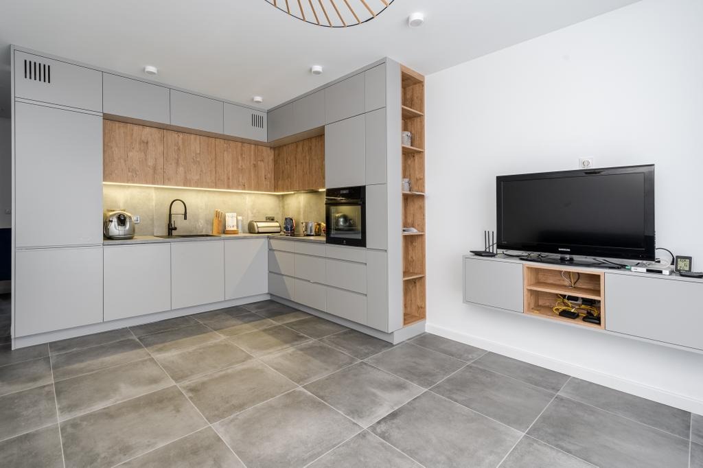 Nowoczesna kuchnia z szarymi szafkami i drewnianymi akcentami, oświetleniem LED pod szafkami wiszącymi i płytkami podłogowymi w odcieniach szarości oraz ścianką RTV z telewizorem.