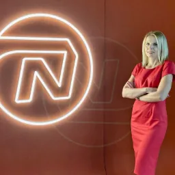 Portret kobiety w czerwonej sukience stojącej na tle ściany z neonowym logo firmy.