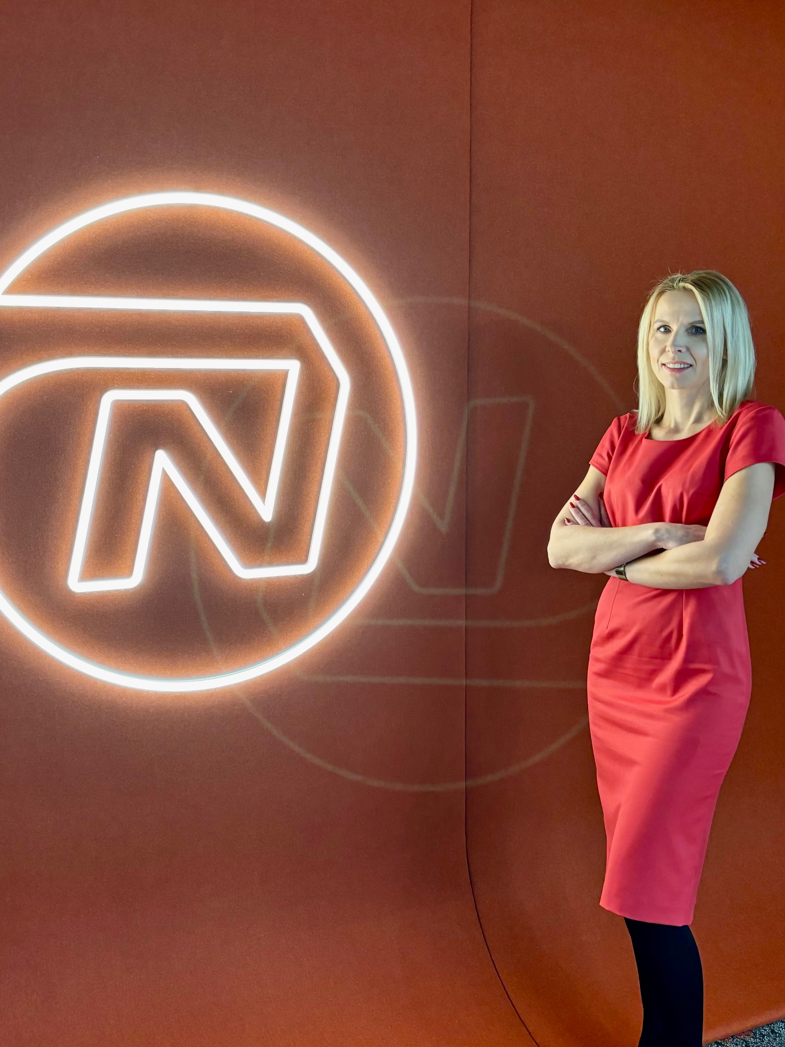 Portret kobiety w czerwonej sukience stojącej na tle ściany z neonowym logo firmy.