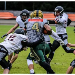 Zawodnicy futbolu amerykańskiego w trakcie meczu na zielonym boisku, widać błoto i dynamiczne ruchy, na koszulce zawodnika z piłką widnieje logo Santander.