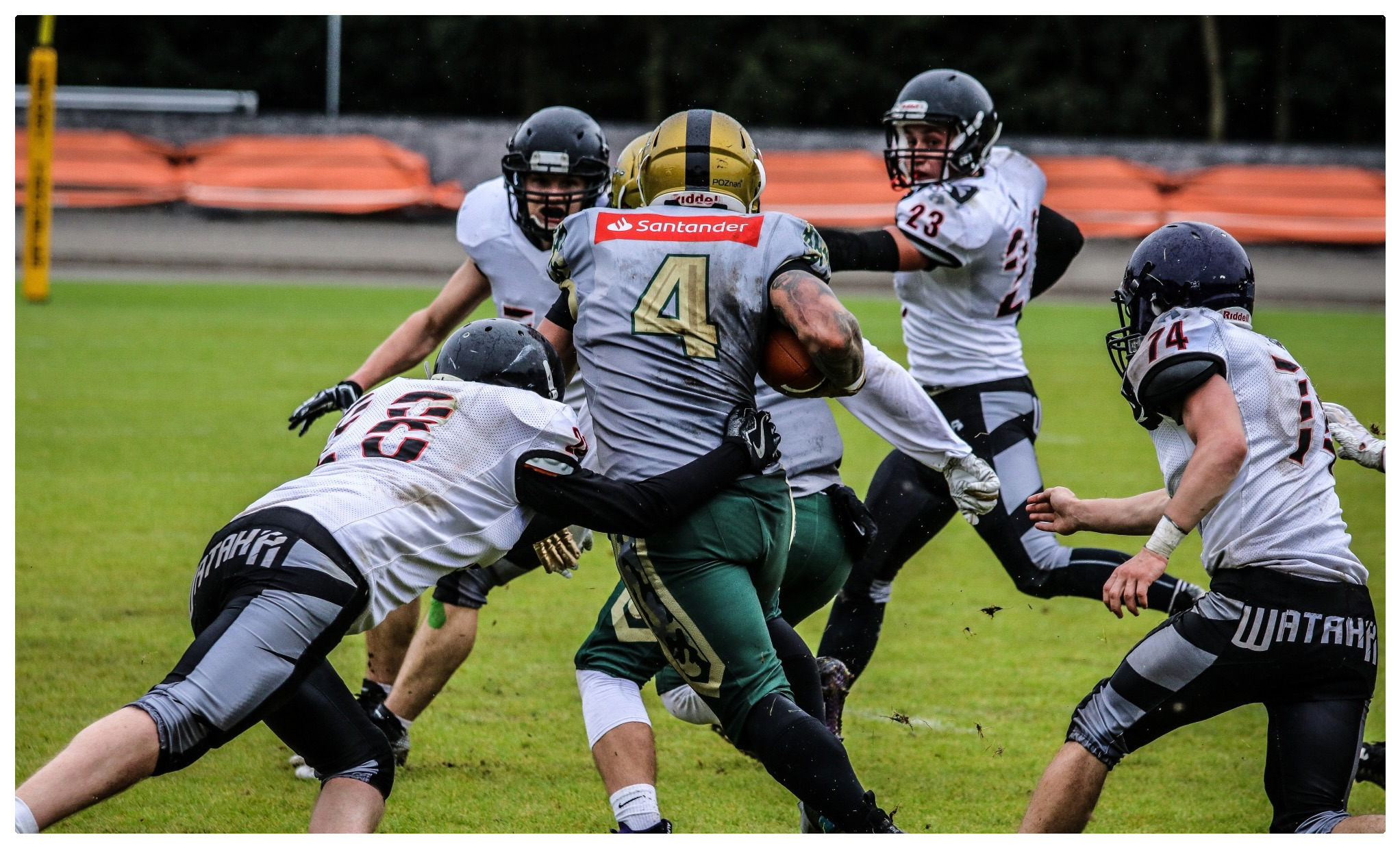 Zawodnicy futbolu amerykańskiego w trakcie meczu na zielonym boisku, widać błoto i dynamiczne ruchy, na koszulce zawodnika z piłką widnieje logo Santander.