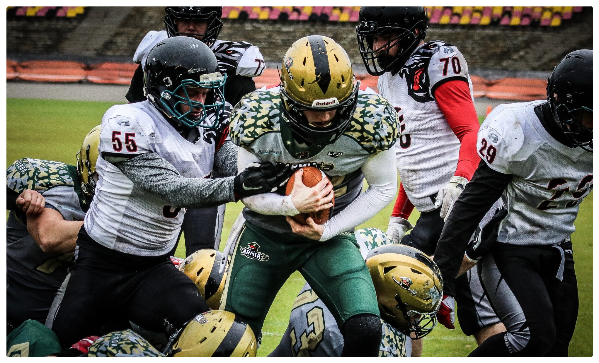 Zawodnicy futbolu amerykańskiego w trakcie zaciętej akcji na boisku, widoczny gracz z piłką w zielono-złotym stroju otoczony przez przeciwników w białych i czarnych uniformach.