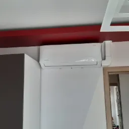 Biały klimatyzator ścienny marki Rotenso zamontowany w nowoczesnym wnętrzu z czerwonym pasem pod sufitem, widoczne elementy zabudowy kuchennej i futryna drzwi.