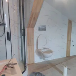 Łazienka w trakcie remontu w Rajczy: widoczna kabina prysznicowa, podwieszana toaleta, biało-marmurowe płytki na ścianach, drewnopodobne panele oraz narzędzia i materiały budowlane.