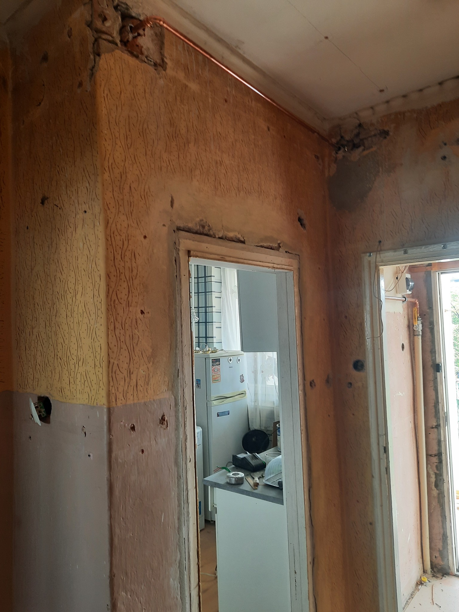 Wnętrze mieszkania w trakcie remontu: ściany z widocznymi śladami po instalacjach, fragment nowej miedzianej rury biegnącej pod sufitem, widok na kuchnię przez otwarte drzwi.