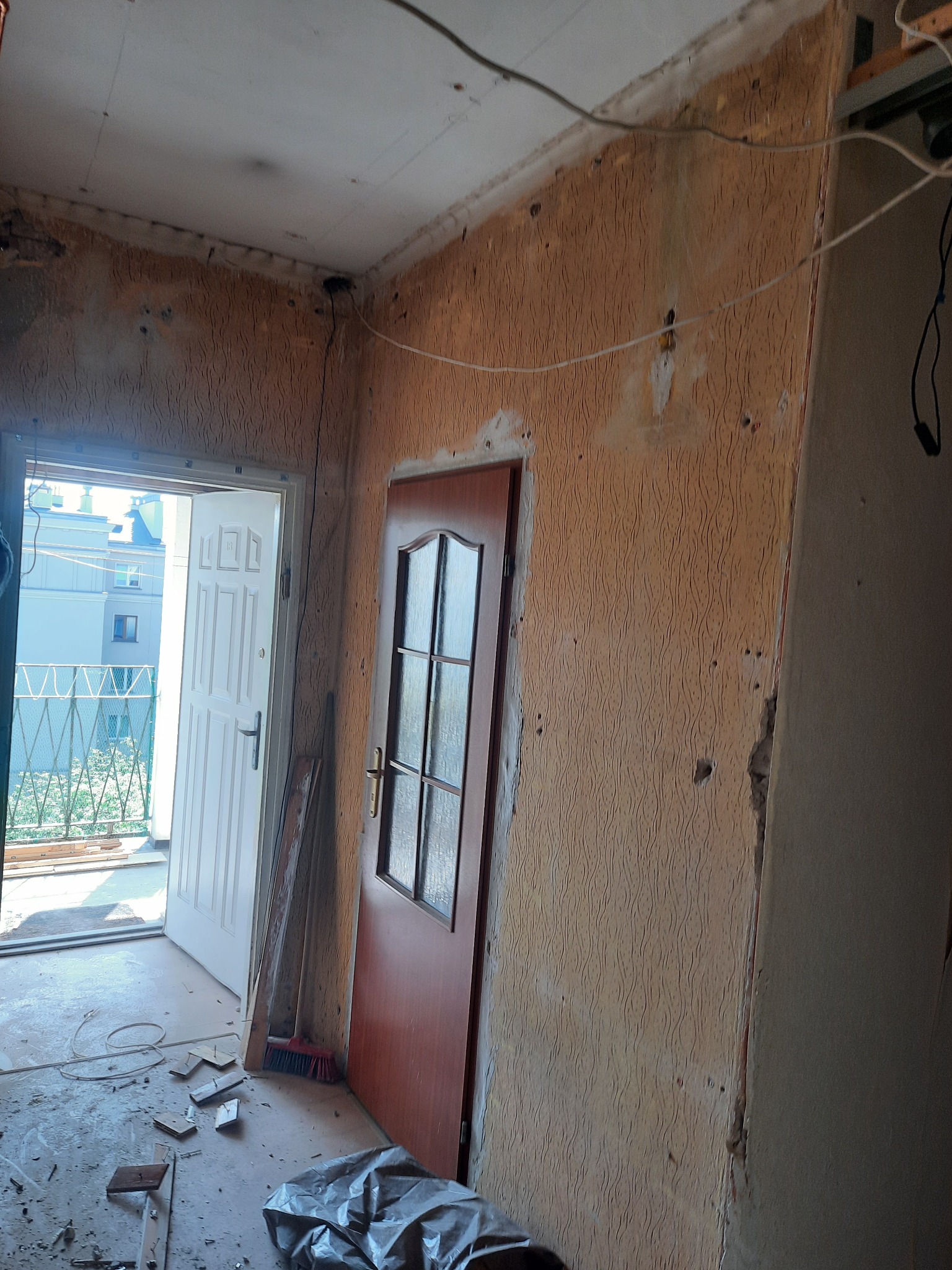 Pokój w trakcie remontu: stara tapeta na ścianach, widoczne ślady po instalacjach, otwarte drzwi balkonowe, gruz i śmieci na podłodze, częściowo wymienione drzwi wewnętrzne.