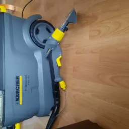 Szaro-żółty odkurzacz piorący Kärcher Professional Puzzi 10/1, leżący na podłodze z paneli.