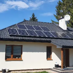 Nowoczesny dom jednorodzinny z ciemną dachówką i panelami słonecznymi na dachu, jasna elewacja, widoczne okna z brązowymi ramami, antena satelitarna na kominie, trawnik w trakcie zagospodarowywania.