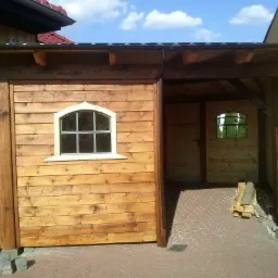 Drewniana konstrukcja z oknem w stylu retro, połączona z budynkiem mieszkalnym, tworząca zadaszony taras z wejściem, wykończona ceglaną kostką brukową.