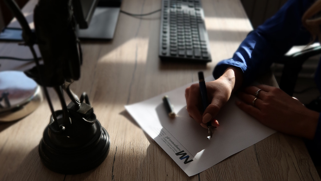 Dłoń kobiety w niebieskiej koszuli, z pierścionkami, pisząca piórem na papierze firmowym na drewnianym biurku, obok statuetka Temidy i klawiatura komputera.