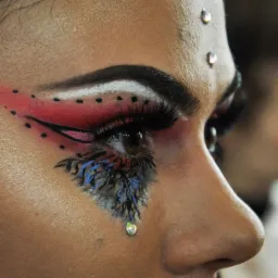 Profil twarzy z artystycznym makijażem: czerwono-czarne kreski, białe akcenty, niebieskie pióra pod okiem i błyszczące kryształki na czole i policzku.