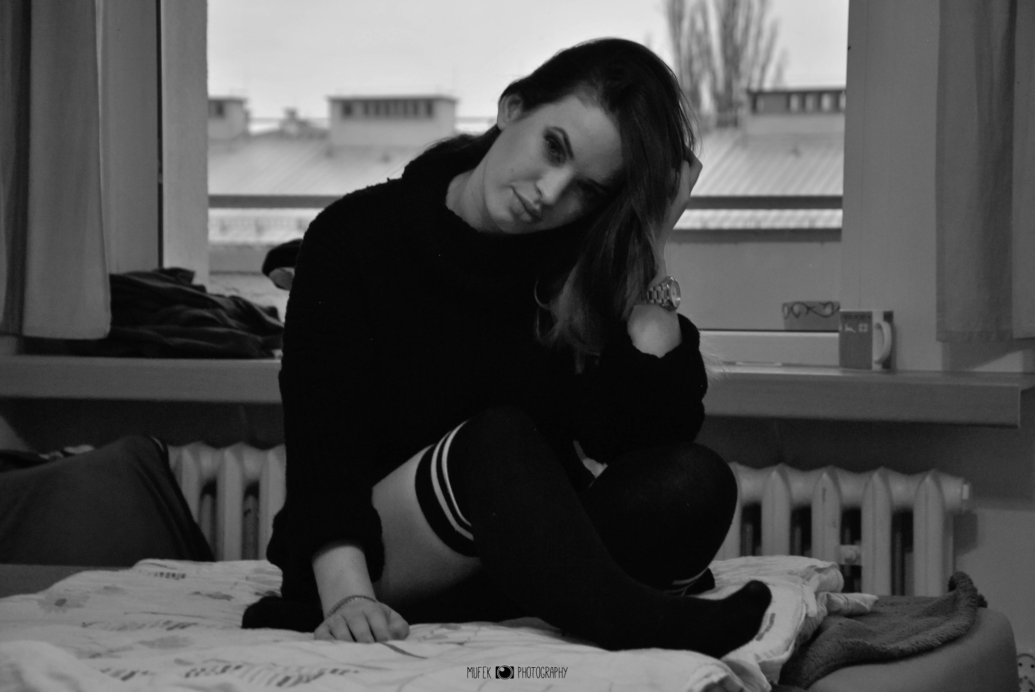 Portret kobiety w czarno-białej tonacji, siedzącej na łóżku przy oknie, w czarnym swetrze i podkolanówkach w paski, z zegarkiem na nadgarstku i dłonią we włosach.