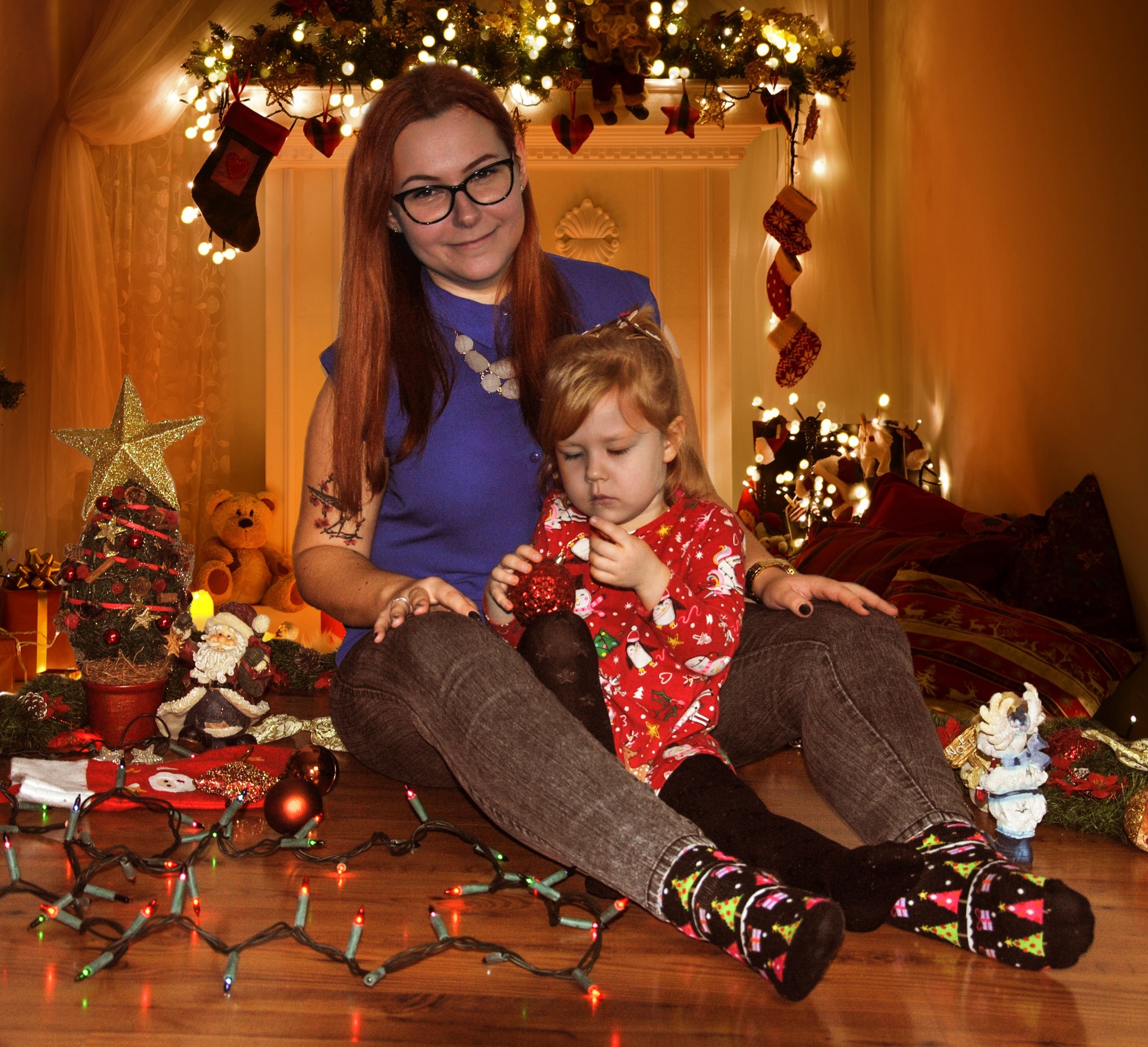 Uśmiechnięta matka w okularach siedzi z córką w świątecznej scenerii z kominkiem, skarpetami, choinką i lampkami, dziewczynka trzyma czerwoną bombkę.