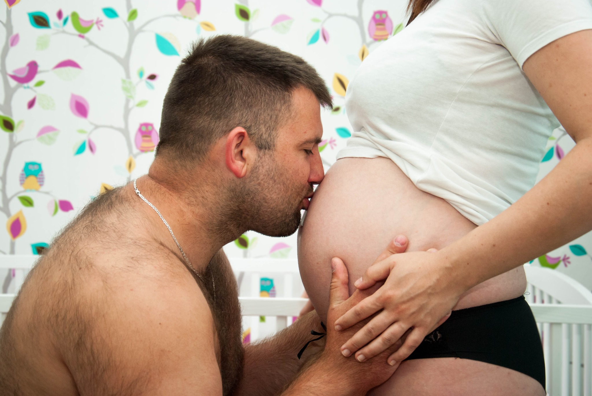Przyszły tata całuje ciążowy brzuszek partnerki na tle ściany w kolorowe sowy i liście, obok białe łóżeczko dziecięce.
