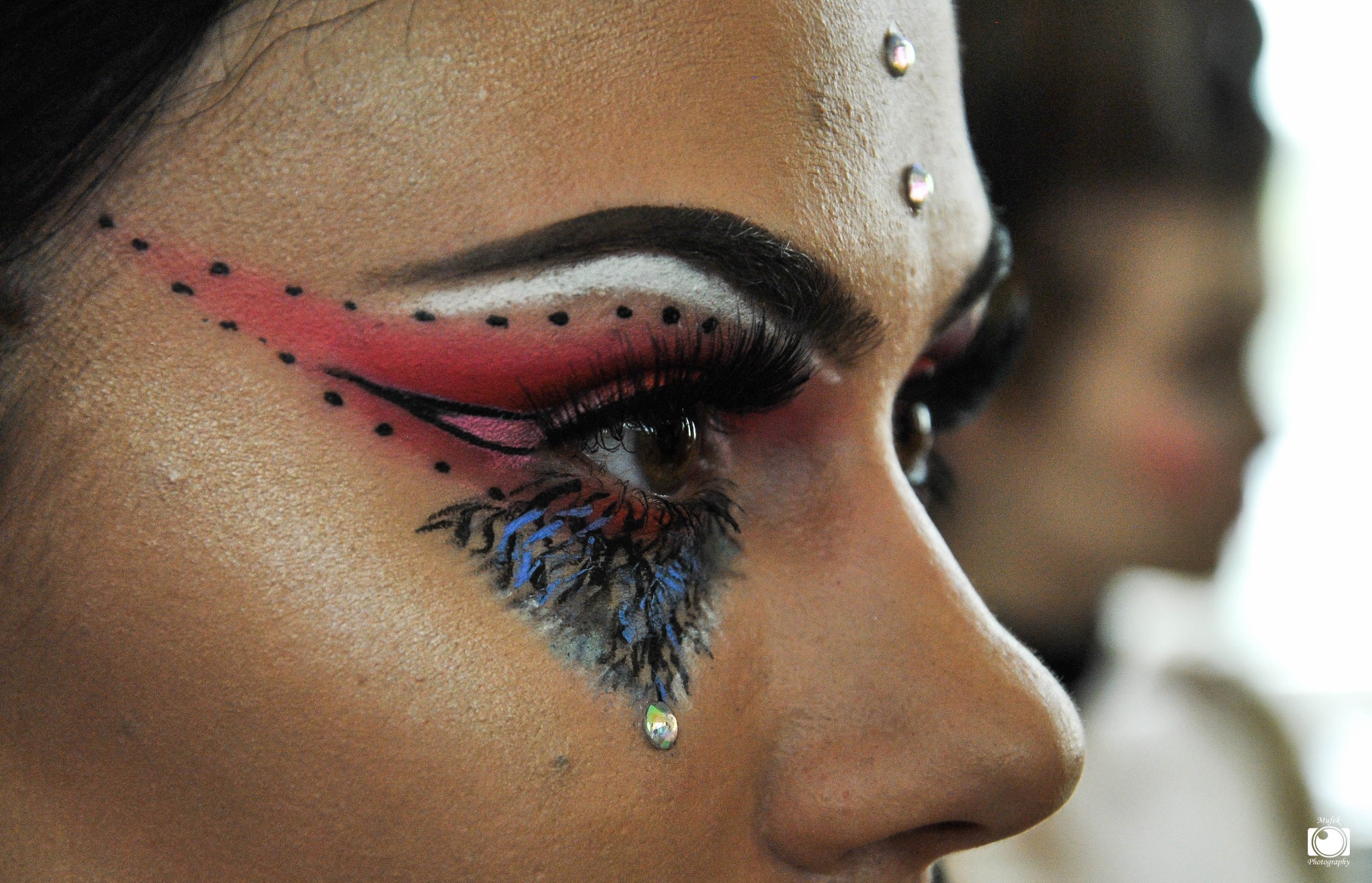 Profil twarzy z artystycznym makijażem: czerwono-czarne kreski, białe akcenty, niebieskie pióra pod okiem i błyszczące kryształki na czole i policzku.