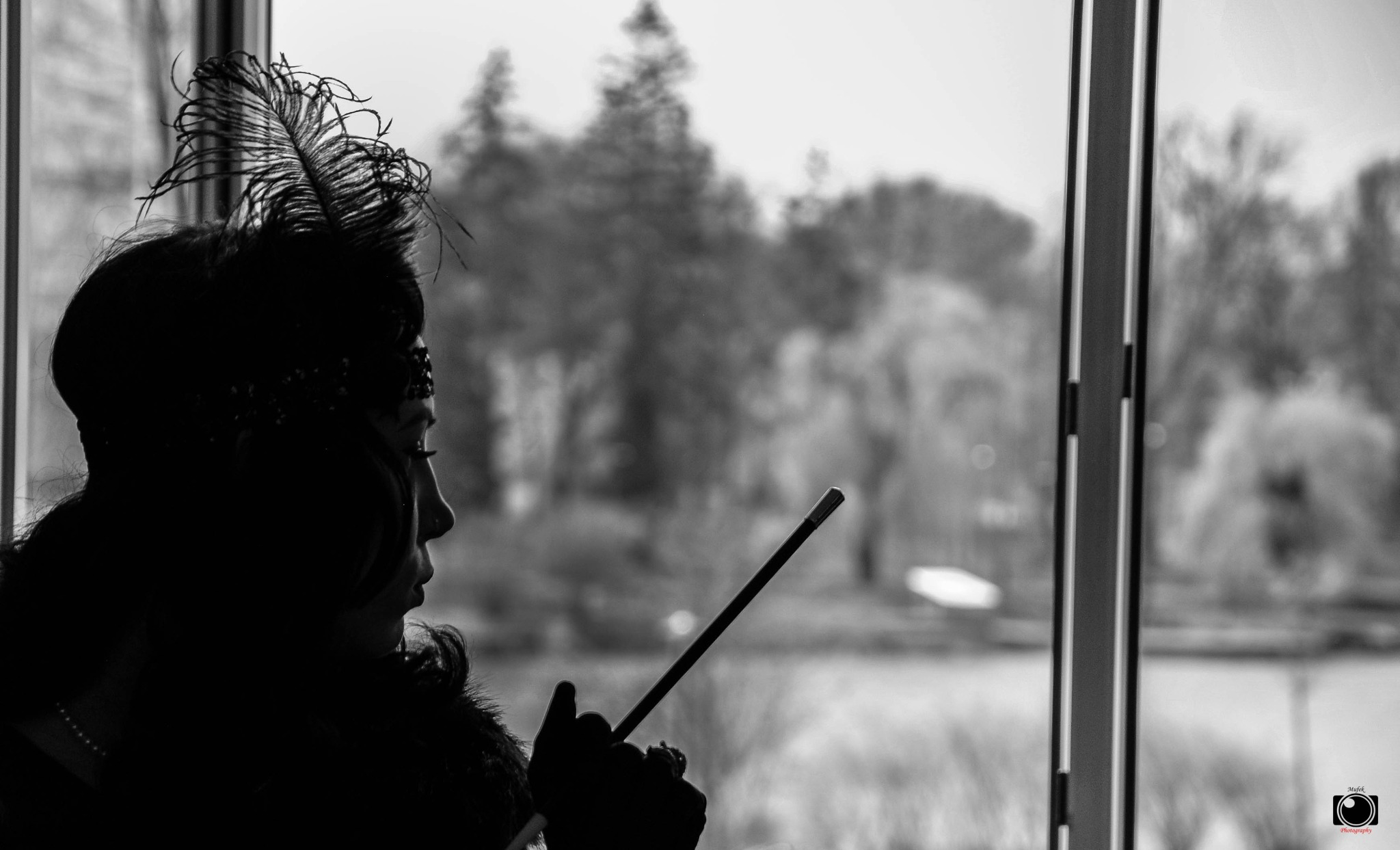 Czarno-białe zdjęcie profilu kobiety w stylu lat 20. z piórem we włosach i długą papierośnicą, stojącej przy oknie z widokiem na zalesiony krajobraz.