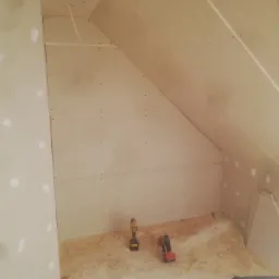 Poddasze w trakcie wykańczania płytami gipsowo-kartonowymi, widoczne dwa wkrętaki akumulatorowe na podłodze z płyt OSB, wystający kabel elektryczny z sufitu.