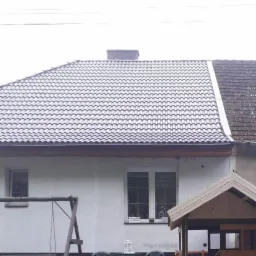 Dom jednorodzinny z nowym, szarym dachem z dachówki ceramicznej, widoczna część starego dachu po prawej stronie, biała elewacja, okno z kwiatami, huśtawka i fragment altanki w ogrodzie.