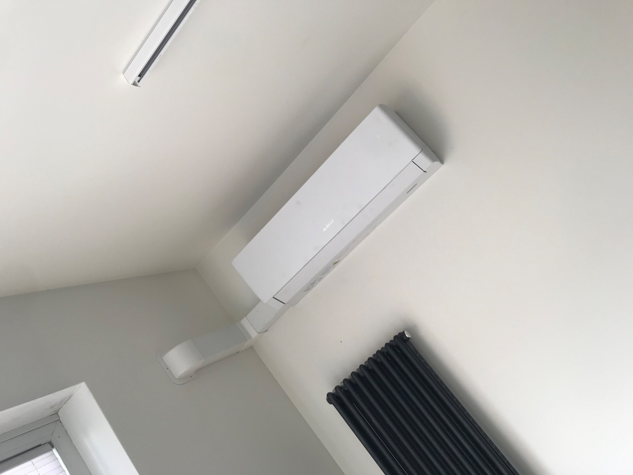 Biały klimatyzator ścienny marki Gree zamontowany w rogu jasnego pomieszczenia, widoczny czarny grzejnik żeberkowy i fragment okna.