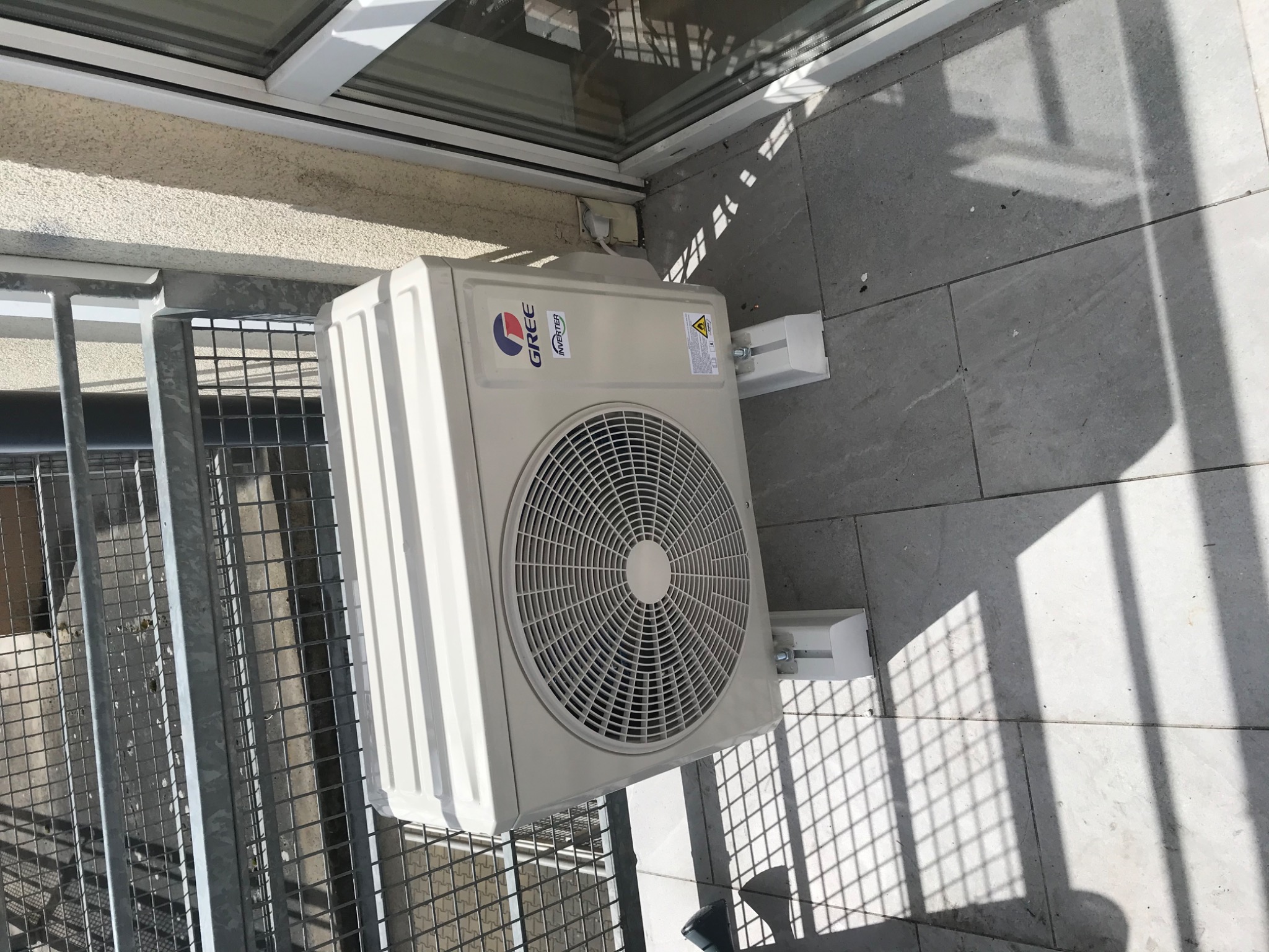 Zewnętrzna jednostka klimatyzacji marki Gree zamontowana na balkonie, widoczna krata balkonu i cień rzucany przez nią na płytki podłogowe.