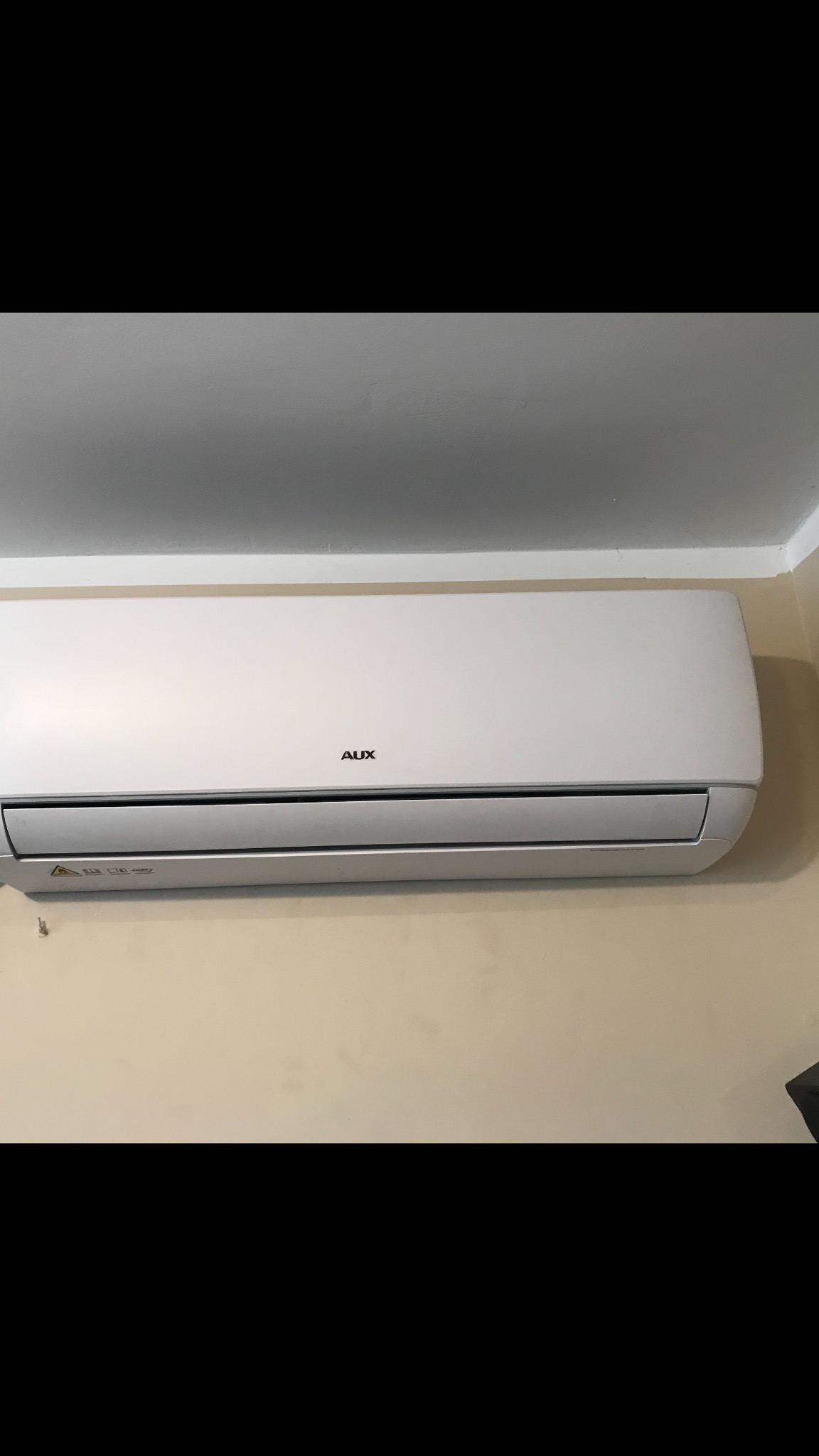 Biały klimatyzator marki AUX zamontowany na beżowej ścianie pod sufitem, widoczne naklejki ostrzegawcze na urządzeniu.