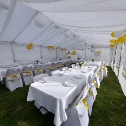 Uroczystość w białym namiocie: długie stoły z białymi obrusami, krzesła z żółtymi wstążkami, białe lampiony i balony w kolorach bieli, złota i srebra.