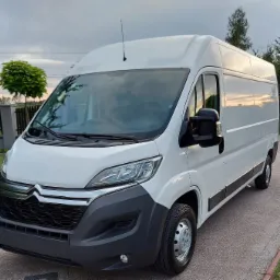 Biały samochód dostawczy Citroen Jumper na betonowym podjeździe, z widocznym odbiciem zachodzącego słońca w bocznej szybie, na tle ogrodzenia i drzew.