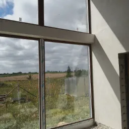 Świeżo położone tynki maszynowe na ścianie wewnętrznej budynku w stanie surowym, widok na pole za oknem z brązową ramą, ślady po pracach wykończeniowych na podłodze.